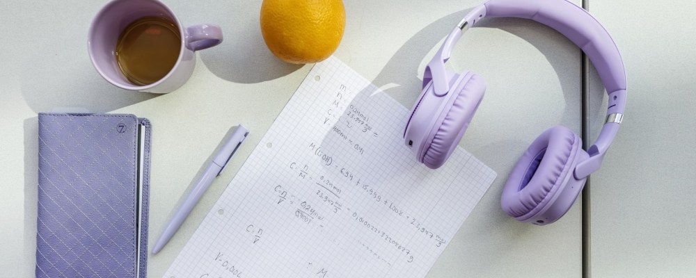 Pöydällä kuulokkeet, kahvikuppi, kalenteri, kynä, appelsiini ja paperi.